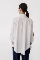 Camisa blanca de algodón con cuello alto con volados, cierre frontal con botones, mangas largas con puños abotonados, bolsillo en el pecho con corazón bordado y parches de crochet en los codos.