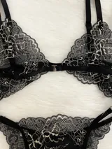 Conjunto de lencería de encaje negro con blanco, compuesto por un corpiño triangular con breteles regulables y cierre de gancho delantero, y una tanga regulable.