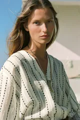 Blusa color crema con rayas verticales bordadas en negro. Tiene cuello redondo con escote en pico, mangas largas abullonadas y cierre frontal con botones.