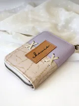Billetera pequeña de cuero color lila y beige con cierre metálico y detalle de cordones cruzados en el frente.
