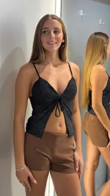 Top negro con brillos, escote en V y tirantes finos. Presenta un detalle de nudo frontal que crea una abertura en la parte inferior.