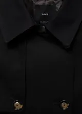 Trench coat negro con cuello de solapa, doble botonadura, trabillas en los puños, cinturón ajustable y abertura posterior en el bajo.