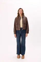 Campera corta color beige claro, estilo bomber o trucker, confeccionada en símil cuero suave. Presenta cuello mao, cierre frontal metálico, bolsillos frontales con tapa y botones a presión, y reguladores ajustables en el ruedo.