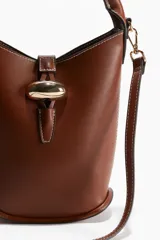 Cartera tipo balde color marrón con asa de mano rígida y correa bandolera ajustable y extraíble. Cierre con trabilla y adorno metálico dorado.