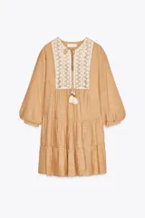 Vestido corto color beige con cuello redondo, cordón con borlas y manga larga. Presenta bordados combinados a contraste en la pechera, cierre frontal con botones ocultos, forro interior y bajo con volantes.