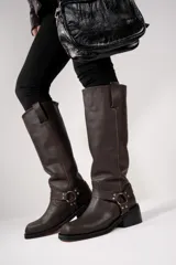 Botas de cuero color marrón oscuro, de caña alta, con taco cuadrado y correa con hebilla metálica alrededor del tobillo.