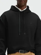 Buzo negro con capucha y cordones ajustables, corte holgado y pequeño logo bordado en el pecho. Se lleva sobre una camisa blanca de cuello.