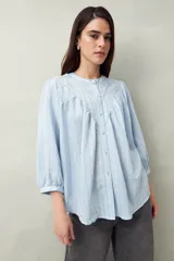 Camisa celeste a rayas verticales, de corte amplio y mangas 3/4. Presenta cuello mao, cierre frontal con botones irregulares y detalle de pespuntes y pinzas en el canesú.