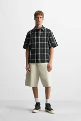 Camisa de hombre relaxed fit con estampado de cuadros blancos sobre fondo negro, cuello solapa, manga corta y cierre frontal con botones.