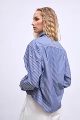 Camisa de mangas largas a rayas verticales azules y blancas, con cuello clásico y cierre frontal con botones. Presenta apliques de strass y ojalillos metálicos.