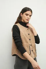 Chaleco color beige de corderito con cuello redondo, cierre frontal con botones a presión y dos bolsillos delanteros con ribete de cuero sintético negro.