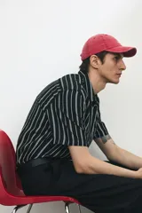 Camisa de manga corta con rayas verticales blancas sobre fondo negro, cuello inglés, tapeta con botones y bolsillo de parche en el pecho. Corte recto y holgado.