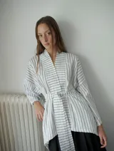 Kimono blanco con rayas verticales negras, confeccionado en tela camisera. Presenta cuello mao, mangas anchas 3/4, bolsillos laterales invisibles y cinturón ajustable desmontable.