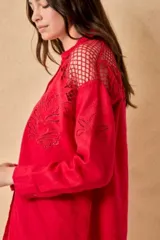Camisa roja de lino con mangas largas, cuello camisero, cierre frontal con botones forrados y bordados florales. Presenta recortes de red calada en los hombros y bordados Millie en las mangas.