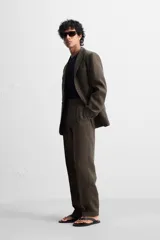 Pantalón de vestir color marrón oscuro, confeccionado en lino italiano. Presenta corte regular fit, cintura con pliegues, bolsillos laterales y traseros ribeteados, y terminación con dobladillo. Cierre frontal con cremallera y botón.