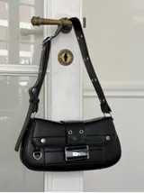 Cartera de hombro negra de cuero sintético, con solapa frontal decorada con una hebilla rectangular metálica plateada y detalles de tachas redondas. Posee correa ajustable para llevar al hombro.
