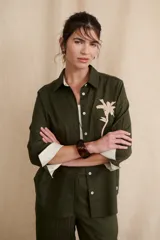 Camisa verde militar de manga larga con puños color crema, cuello clásico y cierre frontal con botones. Tiene un bolsillo en el pecho con bordado de una palmera color crema.