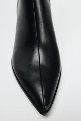 Botín negro de cuero sintético con punta fina y tacón kitten. Presenta una tira con hebilla metálica lateral y cierre mediante cremallera lateral. Tacón de 5,5 cm.