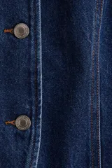 Chaleco largo de denim azul, con cuello redondo, cierre frontal con botones metálicos hasta la cintura y abertura inferior. Presenta bolsillos delanteros insertados y costuras decorativas.