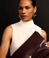 Blusa color crema sin mangas de crepé con cuello alto y lazo por tiras largas de tejido. La prenda tiene cierre por gota con detalle de perla artificial atrás del cuello y frunzidos en la parte delantera.