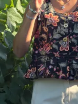 Chaleco negro con estampado floral, cuello redondo y cierre frontal con botones dorados. Presenta corte a la cintura con volantes.