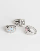 Set de tres anillos plateados con forma de sello. Uno presenta un corazón calado, otro una mariposa celeste y el tercero un moño rosa.