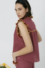 Blusa color mostaza sin mangas, con cuello alto y detalle de volados rojos en cuello, sisas y ruedo. Cierre frontal con botones.
