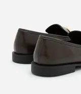 Zapato mocasín de cuero color marrón oscuro con detalle metálico dorado en la parte delantera.
