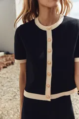 Chaqueta de punto negra con cuello redondo y manga corta. Presenta ribetes en color crema en cuello, mangas y bajo. Cierre frontal con botones dorados.