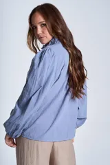 Camisa color beige con volado en el cuello, botones de nácar, tablas decorativas en los hombros y mangas largas con puños elásticos.