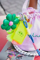 Llavero con cordón trenzado verde neón, detalles en violeta y dijes colgantes: flor verde con centro rosa, borla verde neón y borla fucsia. Incluye tarjetero de plástico rígido color verde neón.