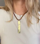 Collar con dije rectangular de bronce texturizado y cordón fino de cuero marrón.