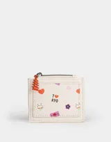 Monedero de cuerina color crudo con cierre superior, bolsillo frontal estampado con motivos japoneses y dije colgante de pagoda roja.