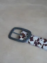 Cinturón de cuero sintético con estampado animal print de vaca en tonos marrones y blancos. Hebilla metálica plateada con logo grabado.