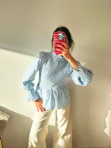 Blusa celeste y blanca a cuadros, con cuello alto, mangas largas abullonadas y elástico en la cintura.
