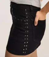 Minifalda de jeans negra, de tiro alto, con cierre tradicional y trabillas en la cintura. Presenta detalles de tachuelas metálicas en los bolsillos delanteros.