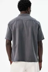 Camisa de manga corta color gris oscuro, confeccionada en mezcla de lino y algodón. Tiene cuello abierto, tapeta lisa, bolsillo de parche en el pecho y bajo recto con aberturas laterales.