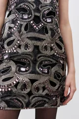 Vestido corto negro con cuello redondo y sin mangas, completamente bordado con mostacillas, piedras y lentejuelas. Tiene corte recto y calados que acompañan el diseño.