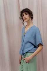 Blusa de lino color azul claro con escote en V cruzado y manga corta. Se ajusta en el centro del busto con un botón redondo de madera o metal con relieve.