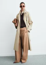Abrigo largo estilo trench coat color beige, con doble botonadura, cuello de solapa ancha, manga larga con puños abotonados y cinturón desmontable. Presenta un diseño recto y oversize.