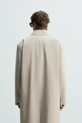 Gabardina color arena de corte recto, con cuello solapa, cierre frontal con botones, mangas largas con puños abotonados, bolsillos laterales y abertura en la parte posterior.