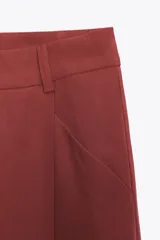Pantalón color ladrillo de tiro medio con trabillas, detalle de pliegues, bolsillos laterales y falsos bolsillos de vivo en la espalda. Cierre frontal con cremallera y botón.