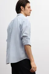 Camisa de hombre de corte holgado, confeccionada en tela de sirsaca a rayas verticales celestes y blancas. Tiene cuello inglés, tapeta clásica con botones, bolsillo de parche en el pecho, mangas largas con puños abotonados y bajo redondeado.