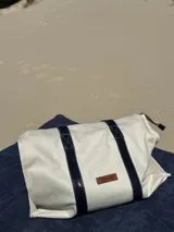 Bolso de lona color blanco con asas de cuero azul marino y etiqueta de cuero con el logo de la marca.