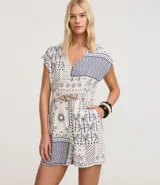 Enterito corto sin mangas de viscolino, con escote en V y estampado tipo patchwork en tonos blanco y azul marino, con motivos tropicales como palmeras y patrones geométricos. Incluye trabillas en la cintura y cinturón de cuerda trenzada.