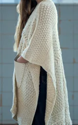 Ruana abierta color crudo, tejida a mano en crochet con lana merino uruguaya. Presenta textura combinada de puntos y trenzas, y bolsillos de cuero vacuno aplicados manualmente.
