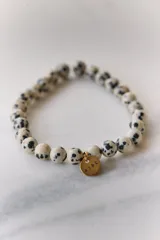 Pulsera elástica de piedras naturales redondas color crema con manchas negras, con dije redondo dorado con la palabra "AIRE" grabada.