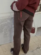 Pantalón de jean marrón de corte recto y tiro medio, con costuras en color naranja y texto estampado en el bolsillo trasero.