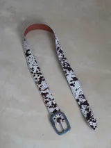 Cinturón de cuero sintético con estampado animal print de vaca en tonos marrones y blancos. Hebilla metálica plateada con logo grabado.