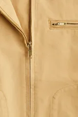 Campera color beige de algodón Oxford con cuello de pana marrón en contraste. Modelo de corte holgado con cierre frontal, hombros caídos, bolsillos laterales ribeteados y bolsillo superior con cierre. Ajuste con botones a presión en los puños y en la parte posterior del bajo.